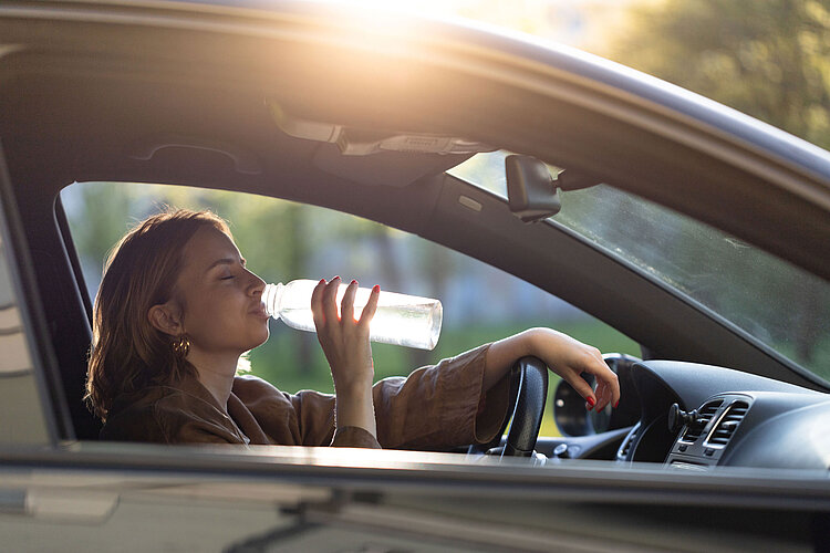 A woman sits at the wheel of a car and takes a sip of water from a bottle.
